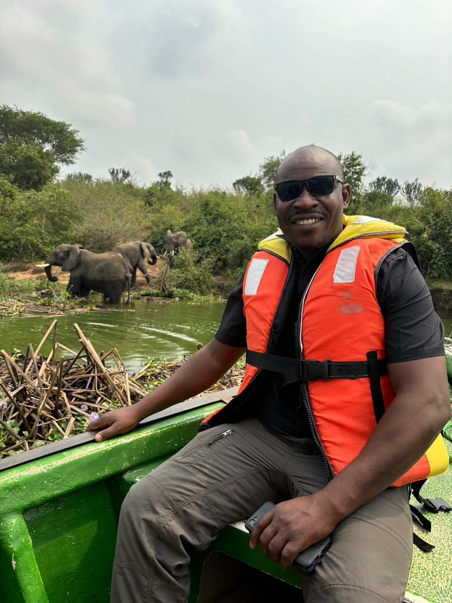 Amanya Godwin on a boat safari on the Nile, with elephants on the riverbank behind him